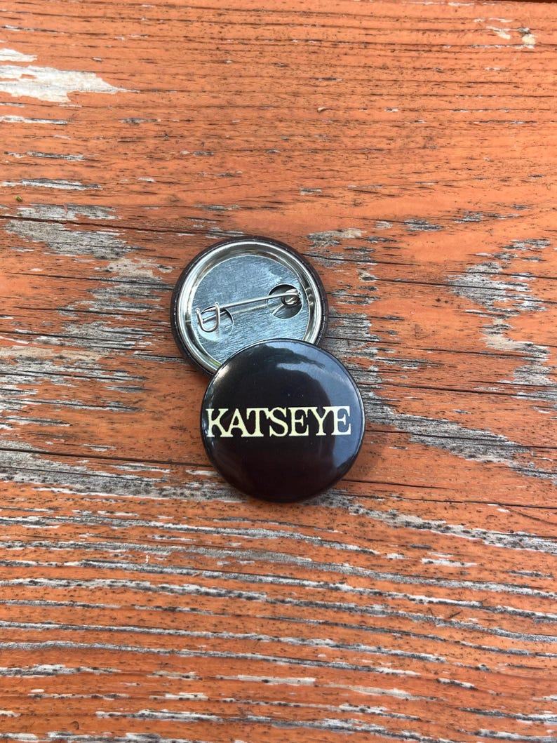 May include: A circular black pin-back button featuring the word KATSEYE printed in a bold, light-colored serif font. The back of the button is metallic silver with a horizontal pin mechanism visible, showing the construction of the accessory.