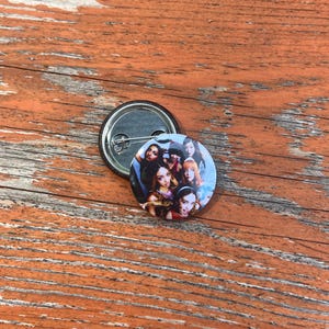 May include: A circular pin-back button featuring a colorful photographic collage of five individuals with diverse hairstyles and expressions. The button has a metallic silver backing with a pin mechanism visible, showcasing the construction of the accessory.