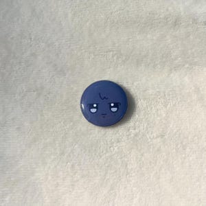 Puede incluir: Un bot&oacute;n redondo azul con una cara de dibujos animados. La cara tiene dos ojos negros, un peque&ntilde;o ce&ntilde;o fruncido y una sola l&iacute;nea por encima de los ojos.