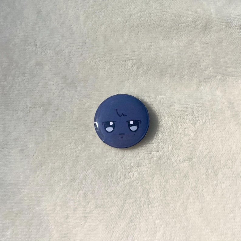 Puede incluir: Un bot&oacute;n redondo azul con una cara de dibujos animados. La cara tiene dos ojos negros, un peque&ntilde;o ce&ntilde;o fruncido y una sola l&iacute;nea por encima de los ojos.