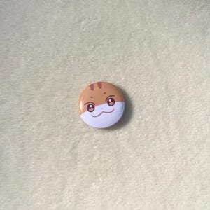 Puede incluir: Un bot&oacute;n redondo con una cara de dibujos animados. La cara es blanca con cejas marrones, mejillas naranjas y ojos negros. El bot&oacute;n es marr&oacute;n por fuera.