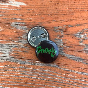 May include: Two circular pin-back buttons. One button shows a black background with the word Gnarly written in a dripping green font. The second button is flipped over to reveal a silver metal back with a horizontal pin mechanism attached.