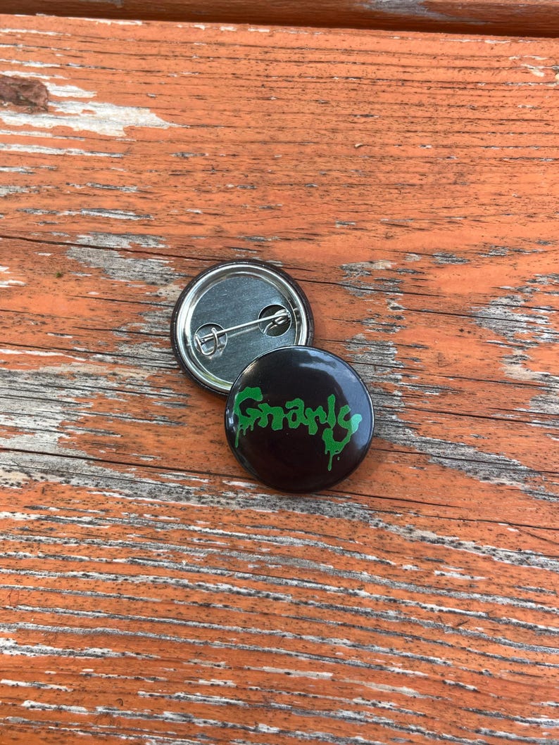 May include: Two circular pin-back buttons. One button shows a black background with the word Gnarly written in a dripping green font. The second button is flipped over to reveal a silver metal back with a horizontal pin mechanism attached.