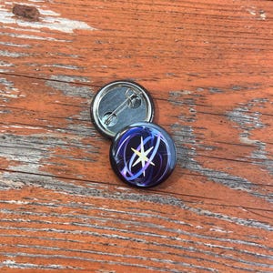 May include: A circular pin-back button featuring a dark purple and black background with swirling light blue and white abstract lines surrounding a central four-pointed yellow and white star icon. The metal back includes a pin mechanism.