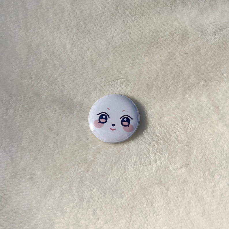 Puede incluir: Bot&oacute;n blanco con una cara de dibujos animados con mejillas rosadas y ojos negros.