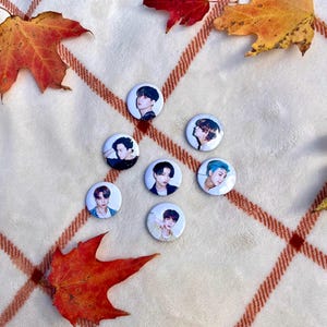 May include: Seven round white button pins with images of different people on them. The pins are on a white and orange plaid blanket with fall leaves.
