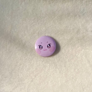 Puede incluir: Un bot&oacute;n morado con una cara de dibujos animados con ojos grandes y mejillas sonrosadas.