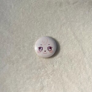 Puede incluir: Un bot&oacute;n blanco con una cara de dibujos animados. La cara tiene dos ojos grandes, una nariz peque&ntilde;a y una boca peque&ntilde;a. La cara est&aacute; sonriendo.
