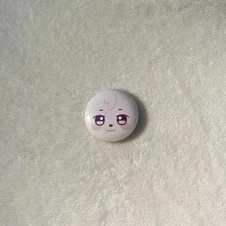 Puede incluir: Un bot&oacute;n blanco con una cara de dibujos animados. La cara tiene dos ojos grandes, una nariz peque&ntilde;a y una boca peque&ntilde;a. La cara est&aacute; sonriendo.