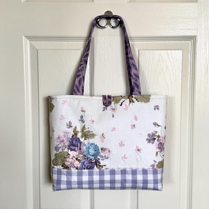 May include: A rectangular fabric tote bag featuring a white background with a floral pattern of blue, purple, and green flowers. The bottom section has a light purple and white gingham check pattern. It includes two purple patterned shoulder straps.