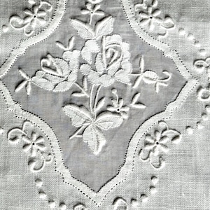 May include: A white textile featuring intricate white embroidery of a central rose motif surrounded by a diamond-shaped border. The fabric displays a visible weave texture, with delicate floral patterns and small eyelet details accentuating the edges.