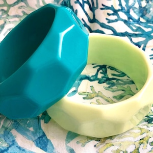 May include: Two chunky bangle bracelets with a faceted geometric surface. One bracelet is a vibrant turquoise blue, and the other is a pale lime green. Both items feature a smooth, glossy finish and a wide, circular band design.