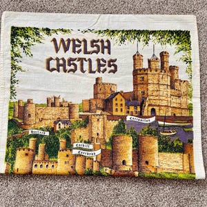 May include: A rectangular white fabric tea towel featuring a colorful illustration of Welsh castles. The text reads WELSH CASTLES, Harlech, Caernarfon, and Cardiff Caerdydd. The design includes stone fortresses, green foliage, and a small boat.