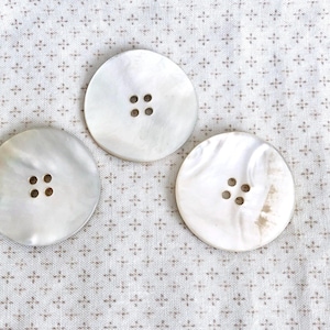 May include: Three large, round, pearlescent white buttons with four holes each. The buttons are arranged on a light beige fabric with a repeating pattern of small, dark brown crosses.