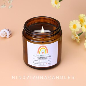Può includere: Un barattolo di candela in vetro marrone con un'etichetta bianca che recita "GET WELL SOON" con un disegno arcobaleno. La candela è etichettata "BLUE WIND CHIME" e ha un tempo di combustione di 16 ore.
