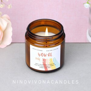 Può includere: Una candela profumata alla lavanda accesa in un barattolo di vetro marrone. L'etichetta recita "You're my Hero" con un messaggio sottostante. La candela ha un tempo di combustione di 16 ore. Il marchio "NINOVIVONACANDLES" è in basso.