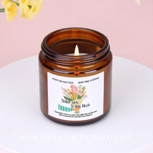 May include: An amber glass candle with a lit wick, displaying a label with floral art and the text "Thank You So Very Much". The candle is labelled "Scent: Sea Salt Sage" and "Burn Time: 16 Hours". The candle sits on a white surface.