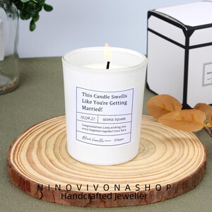 May include: A white candle in a glass jar with a lit wick, on a wooden slice. The candle has a label with the text "This Candle Smells Like You're Getting Married!" and a date. The candle is scented Black Vanilla and has a 15-hour burn time.