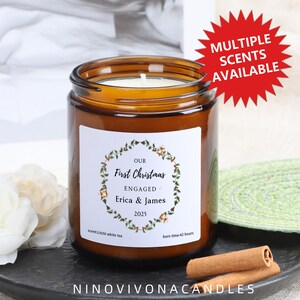 Può includere: Una candela in barattolo di vetro marrone con un'etichetta bianca con la scritta "Our First Christmas Engaged Erica & James 2025". La candela ha uno stoppino bianco ed è circondata da un design a ghirlanda. L'etichetta indica anche "scent: Litchi white tea" e "burn time: 42 hours".