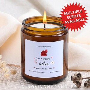 Può includere: Una candela accesa in un barattolo di vetro color ambra con un'etichetta bianca che recita: "A una sorella speciale, Buon Natale". La candela ha una grafica di cappello rosso e il testo "Profumi multipli disponibili". Il profumo della candela è Pera Inglese e Fresia e ha un tempo di combustione di 42 ore.