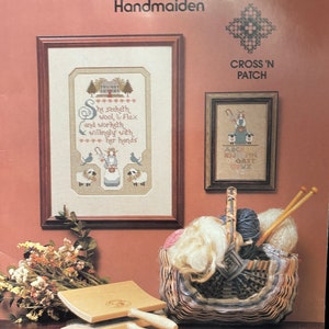 May include: A cross stitch pattern booklet titled "Song of Praise: Handmaiden" with a cross stitch design of a woman spinning wool and a basket of yarn and needles. The text on the pattern reads "She seeketh wool & flax and worketh willingly with her hands".