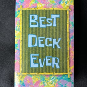 May include: A rectangular box with the text "BEST DECK EVER" in light blue letters. The background is green with vertical lines, framed by a yellow bamboo-like border. The edges feature a floral pattern in pink, purple, and yellow.