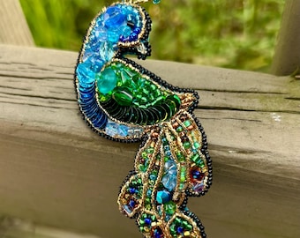 May include: A handcrafted peacock brooch with intricate beadwork. The brooch features a vibrant color palette of blues, greens, and gold, with a detailed design of feathers and a curved body. The brooch is embellished with sequins and beads.