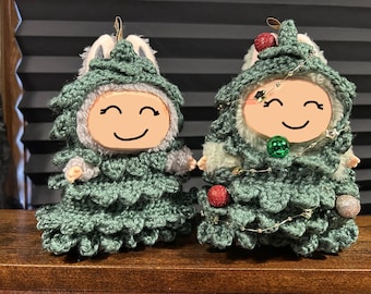 May include: Two handmade Christmas tree ornaments. Each ornament is a doll-like figure with a smiling face, wearing a green crocheted tree costume with white accents. One has red and silver ornaments, the other has a green ornament.