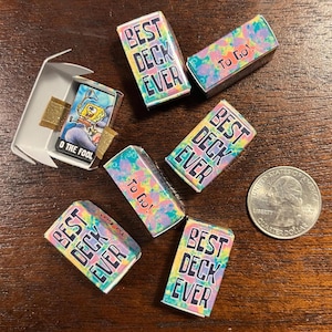 May include: Miniature tarot card decks with colorful tie-dye designs and the text "BEST DECK EVER" and "To Go!" are scattered on a wooden surface. One deck is open, revealing a card with a cartoon character and the text "O THE FOOL". A quarter is also present.