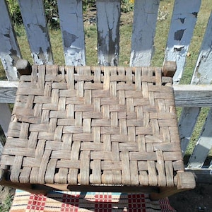 May include: A rustic wooden chair with a woven seat. The seat is made of interwoven strips of wood, creating a textured surface. The chair has a simple design with square legs and a weathered appearance.