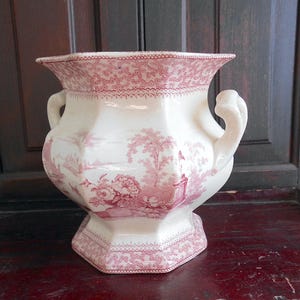 May include: A white and red antique ceramic planter with a detailed landscape design. The planter has a flared rim, two curved handles, and a hexagonal base. The red design includes floral and architectural elements.