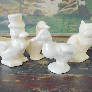 May include: A collection of vintage ceramic figurines in cream and white. The set includes a duck wearing a top hat, two other ducks, and a cat. The figurines are arranged on a dark, textured surface.