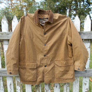 May include: A brown jacket with a corduroy collar and four front pockets hangs on a white picket fence. The jacket has a button-up front and is made of a durable material, suitable for outdoor wear.