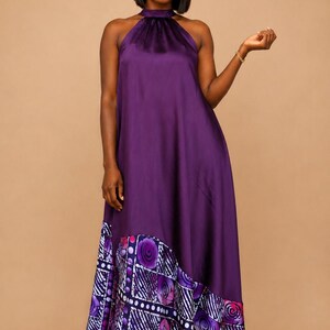 May include: A floor-length, halter-neck dress in a deep purple hue. The dress features a solid, flowing skirt and a patterned hemline with geometric and floral designs in shades of purple, white, and pink.