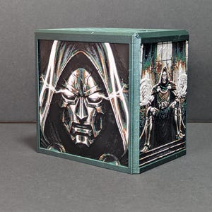 May include: A dark green, rectangular, 3D-printed storage box featuring two distinct graphic panels. The front panel displays a close-up of a metallic, hooded mask with glowing eyes, while the side panel shows a full-body illustration of a cloaked figure.