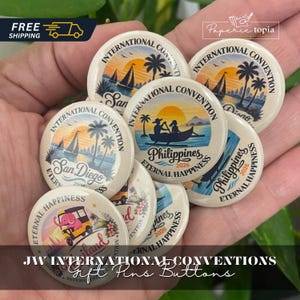 Puede incluir: Una colección de chapas redondas con ilustraciones coloridas de paisajes tropicales, veleros y un elefante rosa en un vehículo. El texto incluye International Convention, San Diego, Philippines, 2026 y Eternal Happiness.