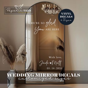 Puede incluir: Un espejo grande y arqueado con texto blanco: "WE'RE SO glad You ARE HERE" y "With love, Jade Matt 05.16.2026". Un círculo azul marino dice "VINYL DECALS 6 Sizes". Las palabras "WEDDING MIRROR DECALS customized sizes" están abajo.