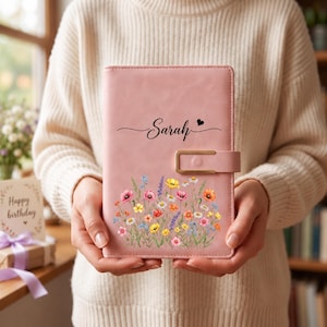 May include: A light pink notebook featuring a gold-toned metal clasp closure. The cover is printed with the name Sarah in black script, a small black heart, and a colourful illustration of various wildflowers in shades of red, yellow, and blue.