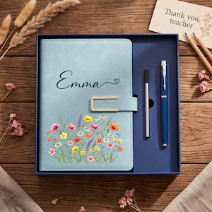 Könnte beinhalten: Ein hellblaues Notizbuch mit dem Namen Emma und einem kleinen Herz in Schwarz, verziert mit einer bunten Wildblumenillustration am unteren Rand, einem goldenen Metallverschluss und einem passenden blauen Stift in einer dunkelblauen Geschenkbox.