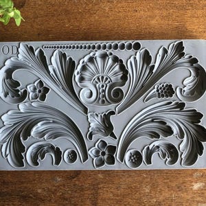 May include: A rectangular gray silicone mold featuring intricate relief designs including large acanthus leaves, a central shell motif, small five-petaled flowers, circular accents, and a row of graduated spheres. The letters IOD are embossed in the top left corner.