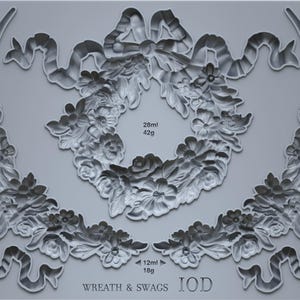May include: A gray silicone mold featuring floral and ribbon designs. The mold includes a central wreath, floral swags, and individual floral and ribbon elements. Measurements are provided in milliliters and grams. The text "WREATH & SWAGS IOD" is visible.