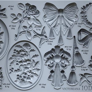 May include: A gray silicone mold featuring various Victorian-style embellishments. Designs include bows, floral arrangements, decorative frames, and a bird. Measurements are indicated in milliliters and grams.