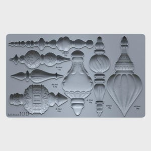 May include: A grey silicone mold featuring various bauble shapes with intricate embossed patterns. The mold includes text reading BAUBLES IOD and specific volume and weight measurements in ml and g next to each cavity, such as 9.3ml 14g, 5.6ml 8.4g, 16ml 24g, 21ml 32g, 12ml 18g, and 24ml 36g.