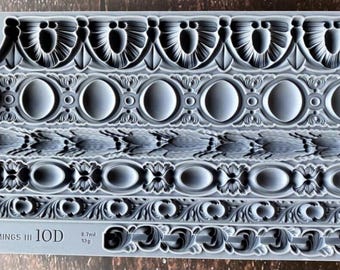Puede incluir: Un molde de silicona gris rectangular que presenta seis filas horizontales distintas de intrincados patrones de borde decorativos. El molde incluye texto en relieve que dice "TRIMMINGS III IOD" y varias medidas de volumen y peso en ml y g.