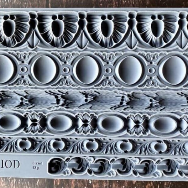 May include: A rectangular grey silicone mold featuring six distinct horizontal rows of intricate decorative border patterns. The mold includes embossed text reading "TRIMMINGS III IOD" and various volume and weight measurements in ml and g.