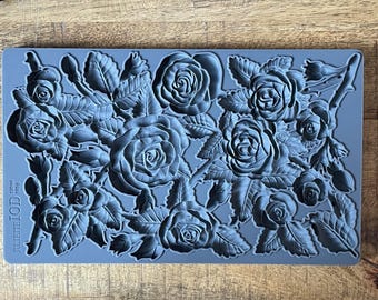 Puede incluir: Un molde rectangular de silicona gris con un patrón en relieve detallado de varias rosas en flor, capullos y hojas dentadas. El texto JULIETTE IOD 130ml 195g está grabado en la esquina inferior izquierda de la superficie flexible.