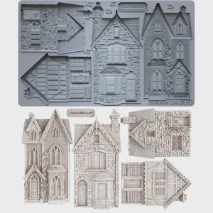 May include: A gray silicone mold for crafting miniature houses, labeled Charlbury Lane IOD, featuring recessed cavities for building facades, roofs, and walls. Below the mold are the finished, light-colored, three-dimensional house structures.