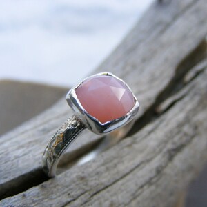 May include: A silver ring with a pink gemstone set in a square bezel. The ring band is decorated with a delicate floral pattern.