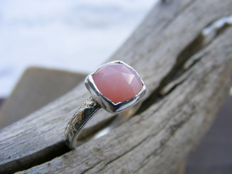 May include: A silver ring with a pink gemstone set in a square bezel. The ring band is decorated with a delicate floral pattern.