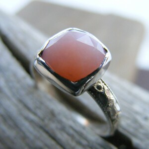 May include: A silver ring with a square, pink gemstone set in a bezel setting. The band is decorated with a floral design.